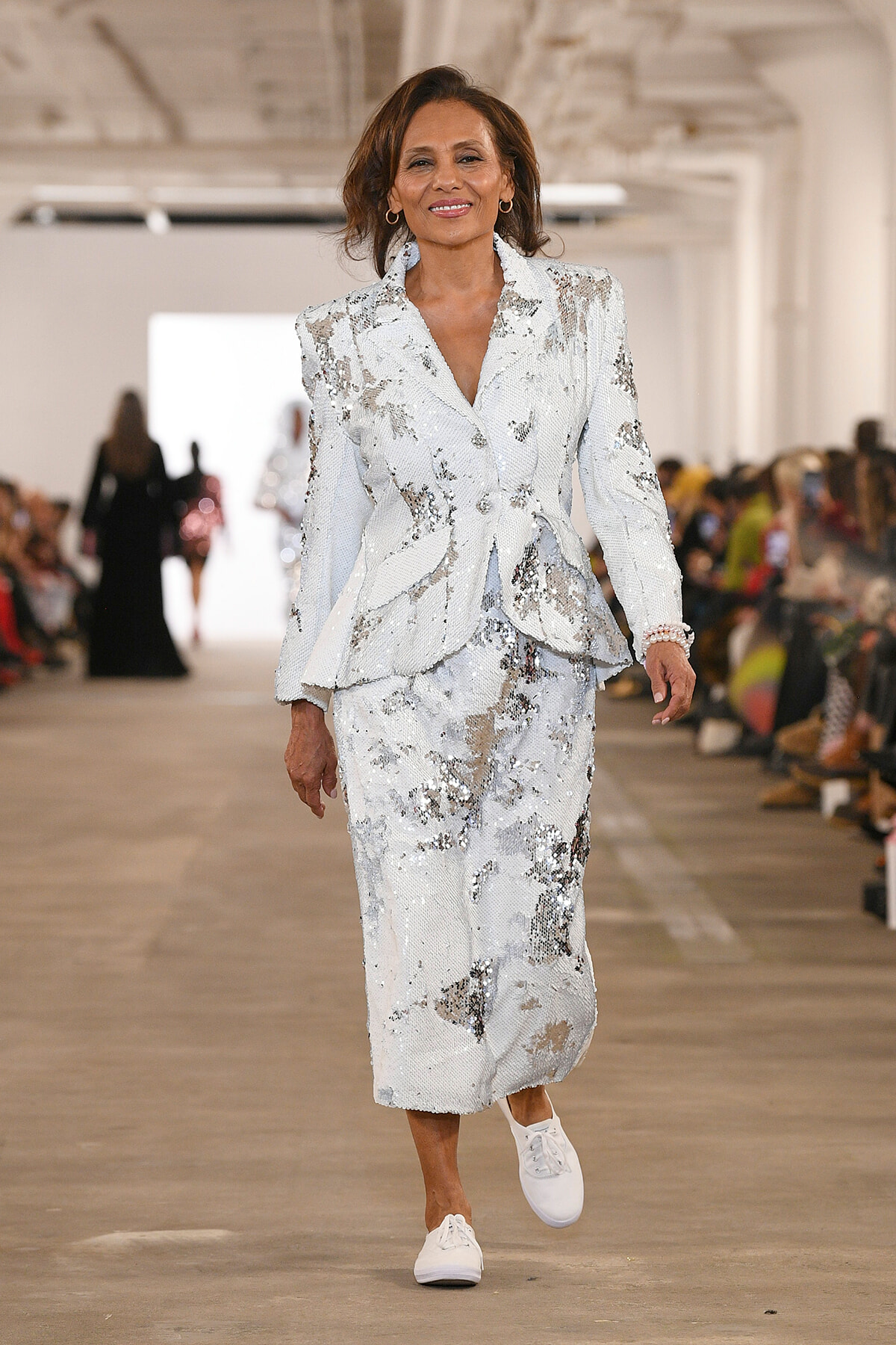 Fashion model walking a runway in a white sequin suit with matching blazer and pants, white sneakers visible, audience in the background.