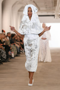 Model walks the runway in a white sequined hooded cape and matching dress, audience blurred in the background.