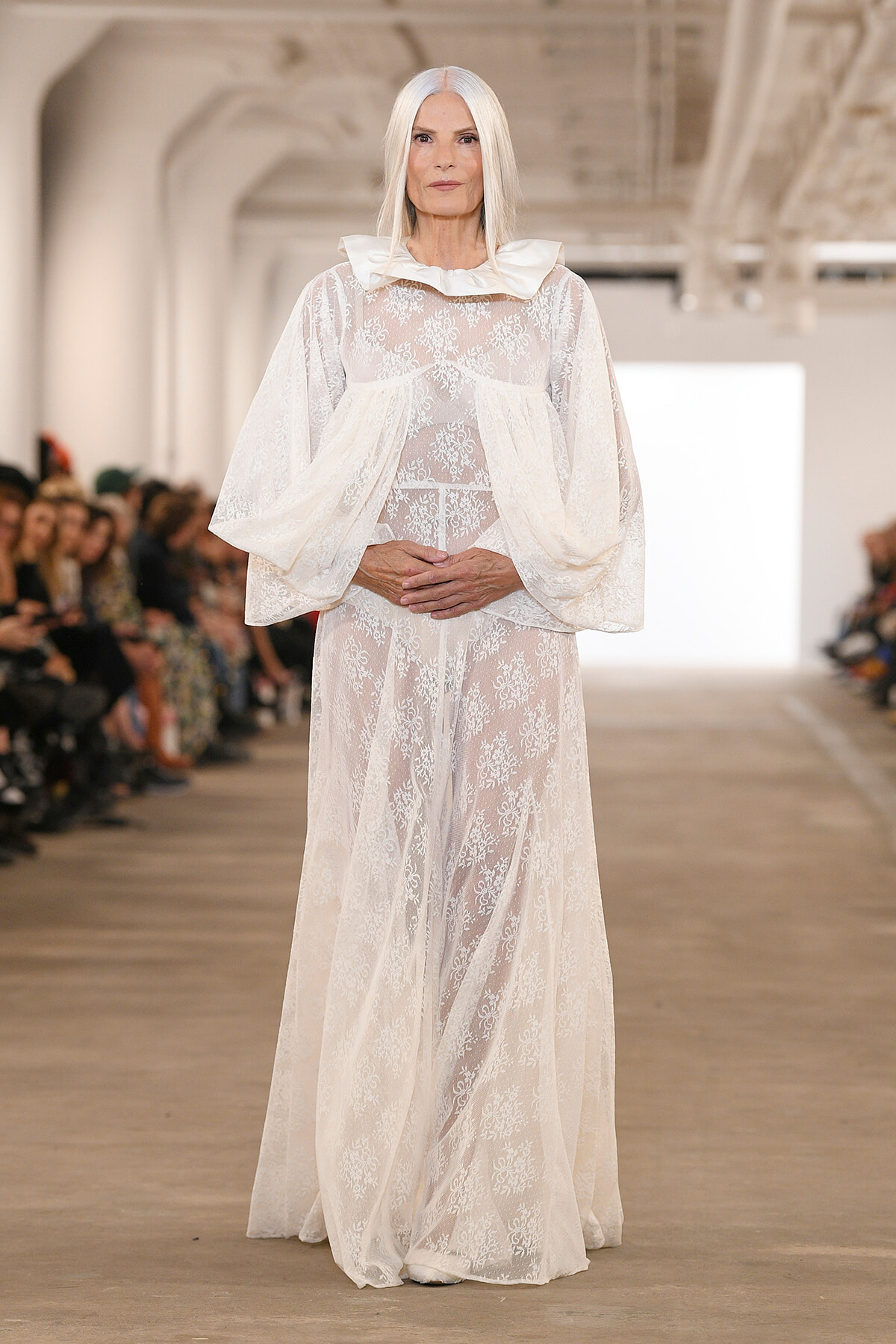 Model in a sheer white lace gown with voluminous sleeves walks a runway; audience blurred in background.