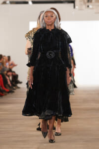 Model walks a runway in a black velvet, ruffled gown with a belt and flower centerpiece; audience blurred in background.