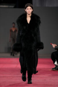 Model walking a red runway in a long black fur coat and boots at a fashion show.