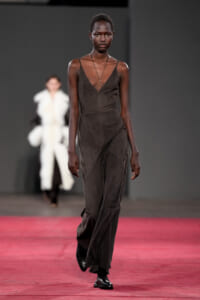 African model walks the runway in a charcoal jumpsuit with thin straps and a pendant necklace, against a gray backdrop on a red carpet.