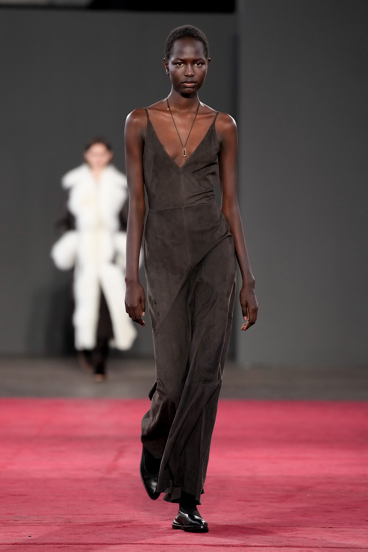 African model walks the runway in a charcoal jumpsuit with thin straps and a pendant necklace, against a gray backdrop on a red carpet.