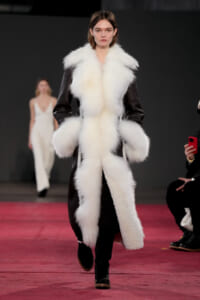Model walking a runway in a black coat with oversized white fur trim at the collar and cuffs.