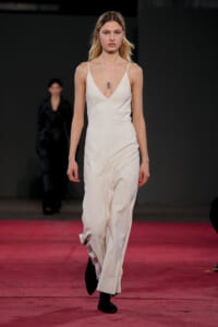 Model walks a runway in a white sleeveless V-neck jumpsuit with a long pendant necklace, on a pink-lit catwalk in a dimly lit venue.