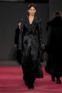 Model walks the runway in a long black leather trench with a belt, clutch in hand, on a red-lit catwalk.
