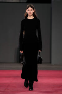 Model walks a runway in a full-length black fuzzy knit dress, carrying a small black handbag. A pink runway contrasts with dark backdrop.