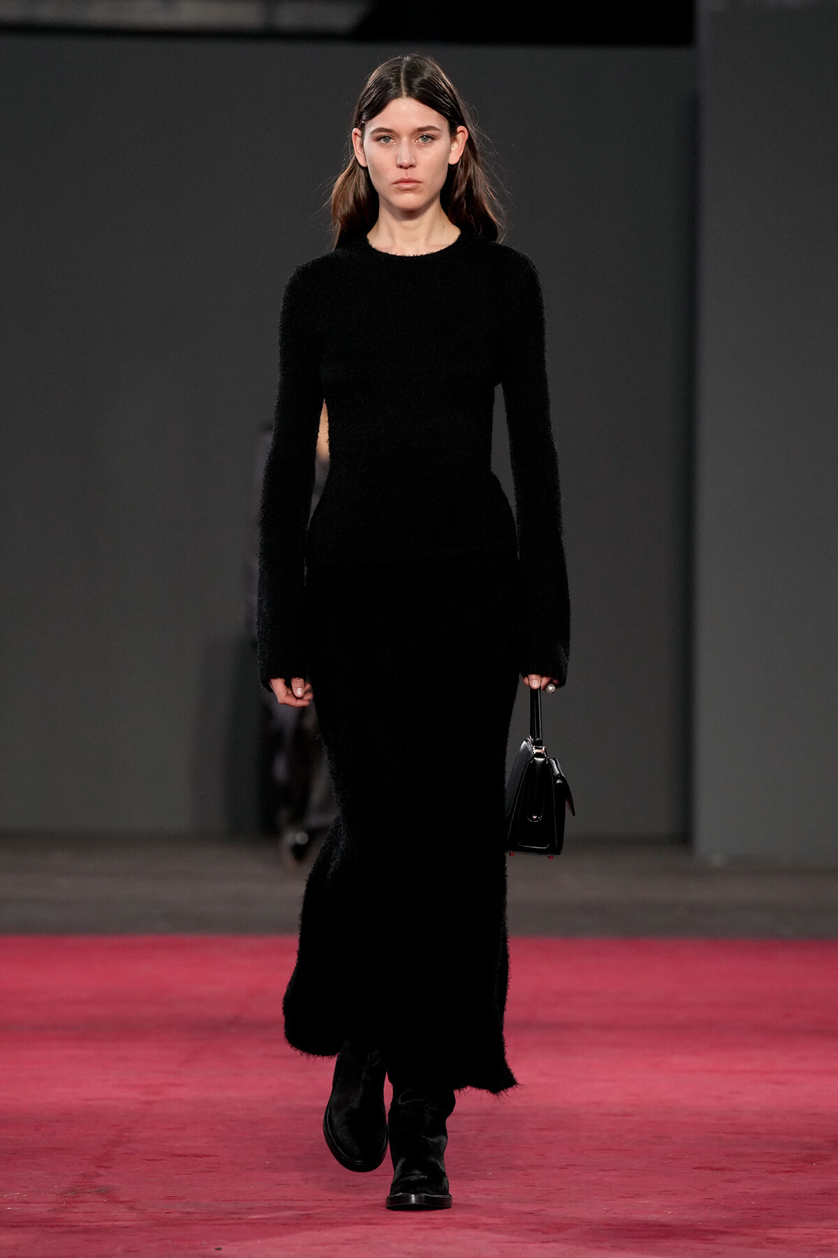 Model walks a runway in a full-length black fuzzy knit dress, carrying a small black handbag. A pink runway contrasts with dark backdrop.