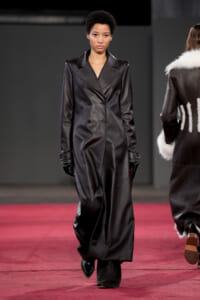 Model walks the runway wearing a long black leather double-breasted coat, gloves, and black boots on a red-carpeted catwalk.