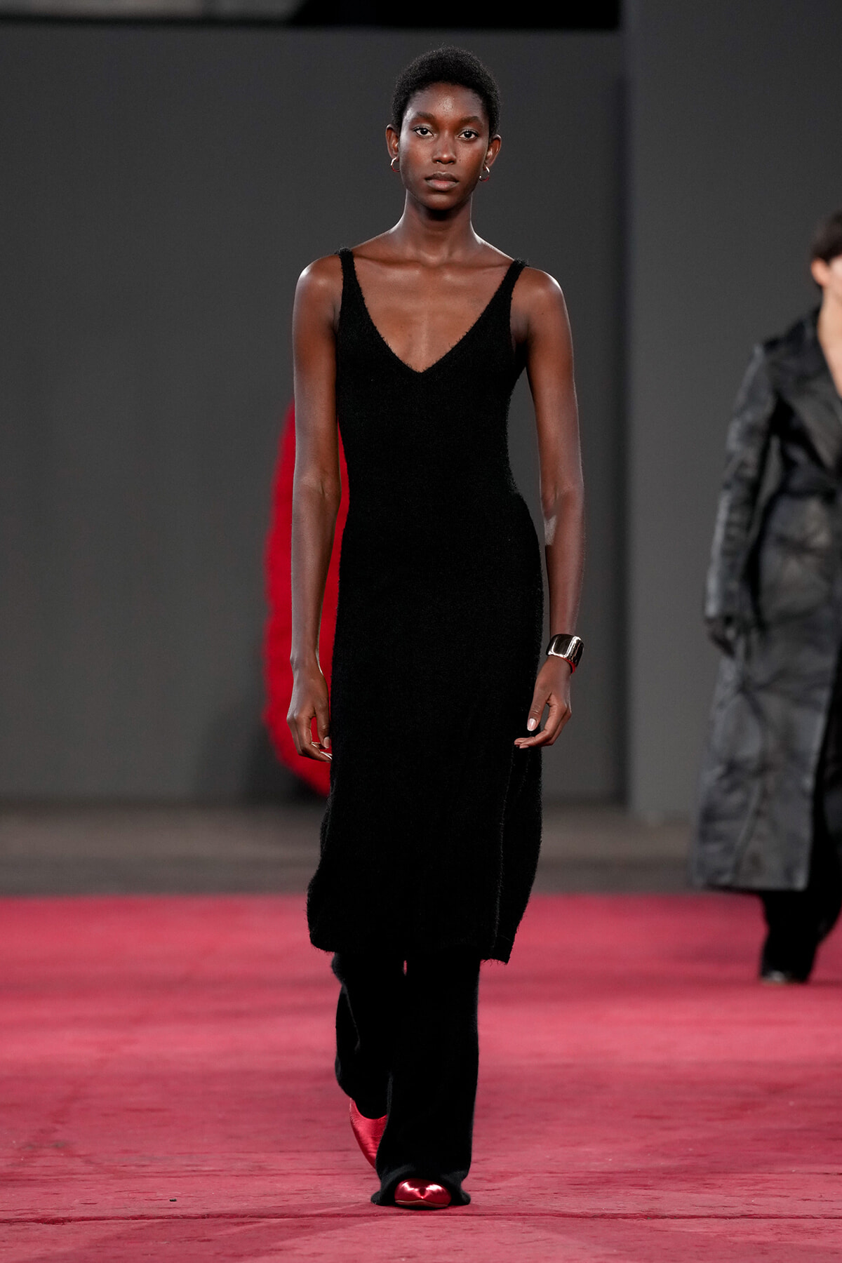 Model walking a fashion runway in a long black V-neck dress with red shoes.