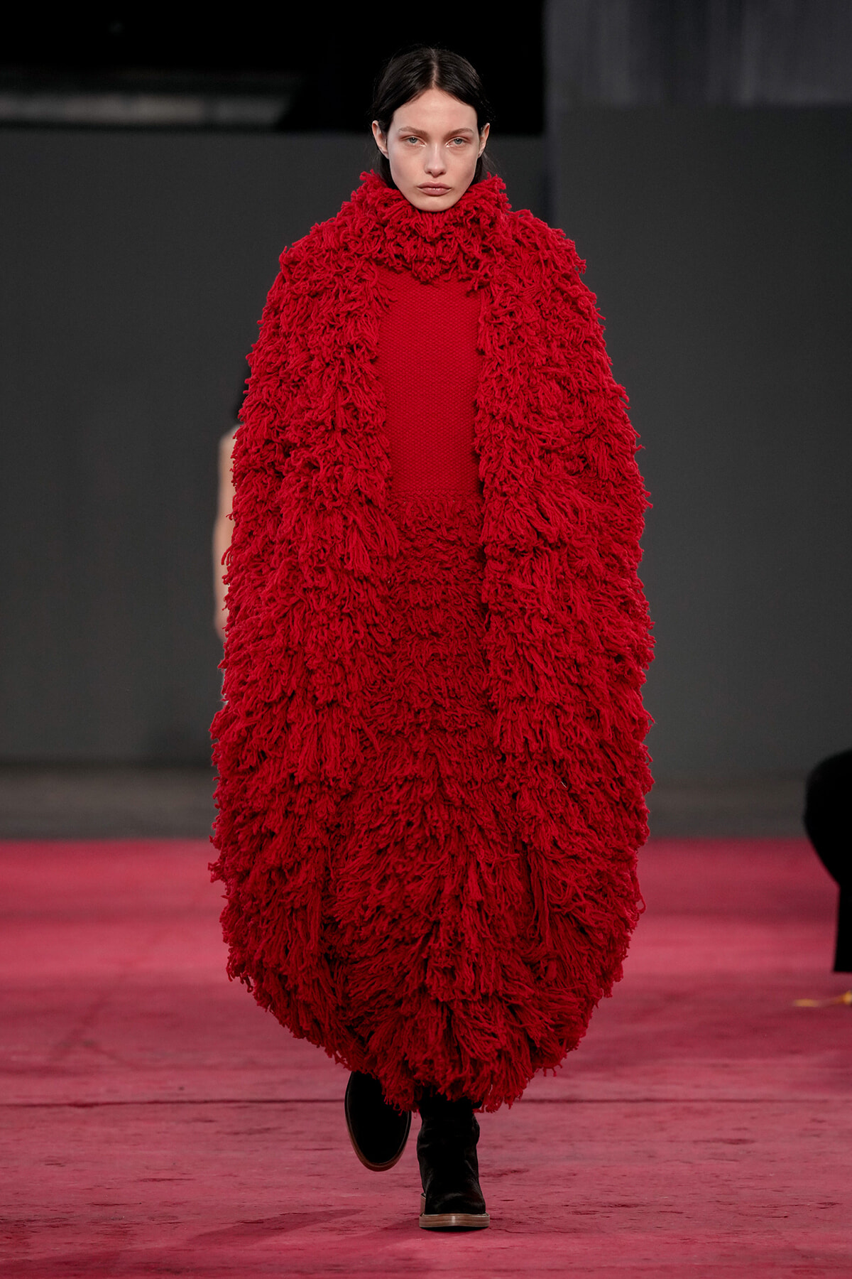 Model walks the runway wearing a voluminous red shaggy coat with a high collar and black boots.