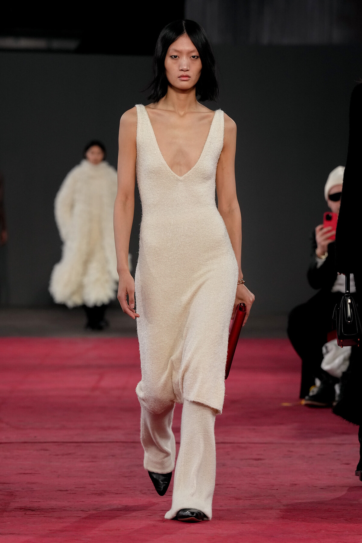 Model walking the runway in a cream textured sleeveless deep-V jumpsuit, holding a red clutch against a dark backdrop on a pink runway.