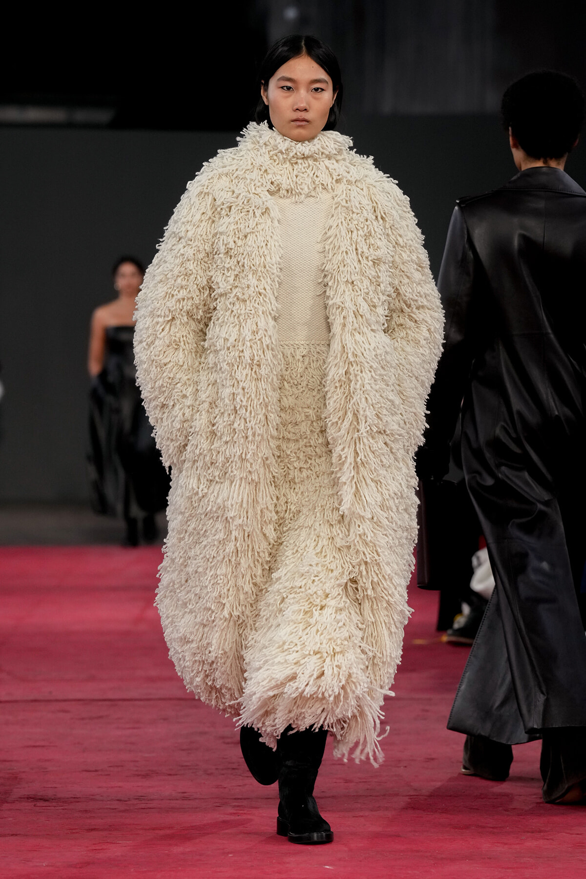 Model walks a runway wearing a voluminous cream shaggy coat and black boots.