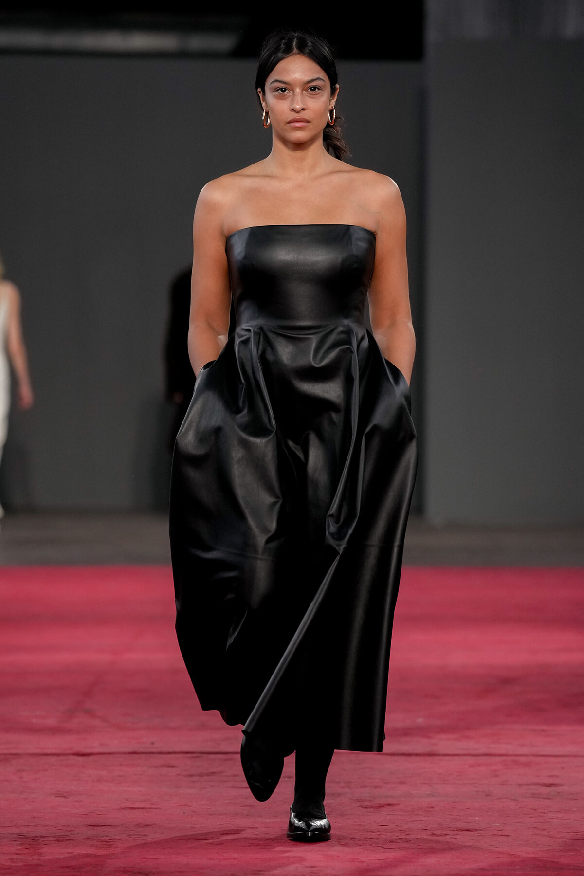 Female model walks the runway in a strapless black satin gown with gold hoop earrings, on a red carpet runway.