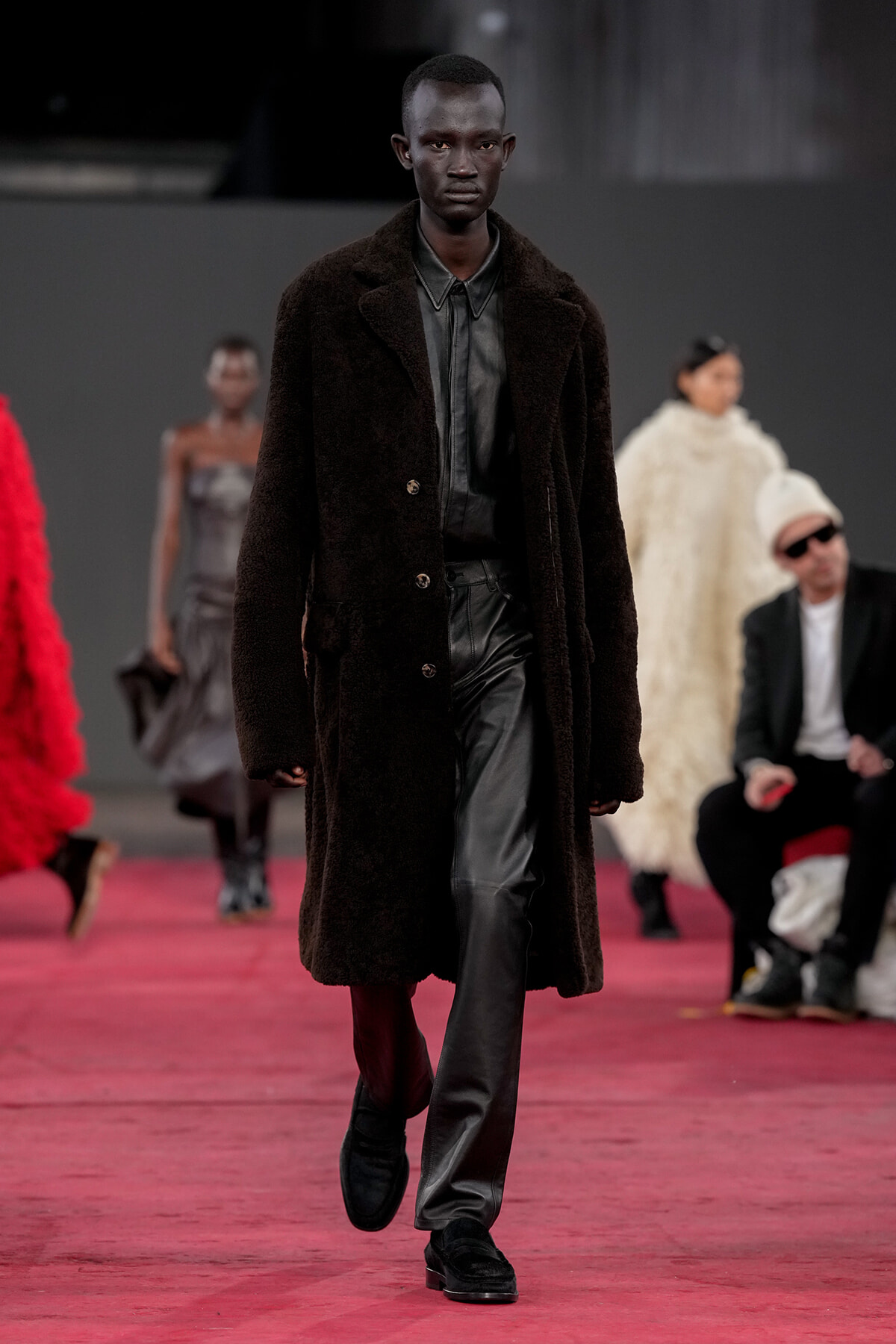 Male runway model wearing a long dark brown shearling coat over black leather pants walks the catwalk on a red/pink carpet with blurred figures in the background.]