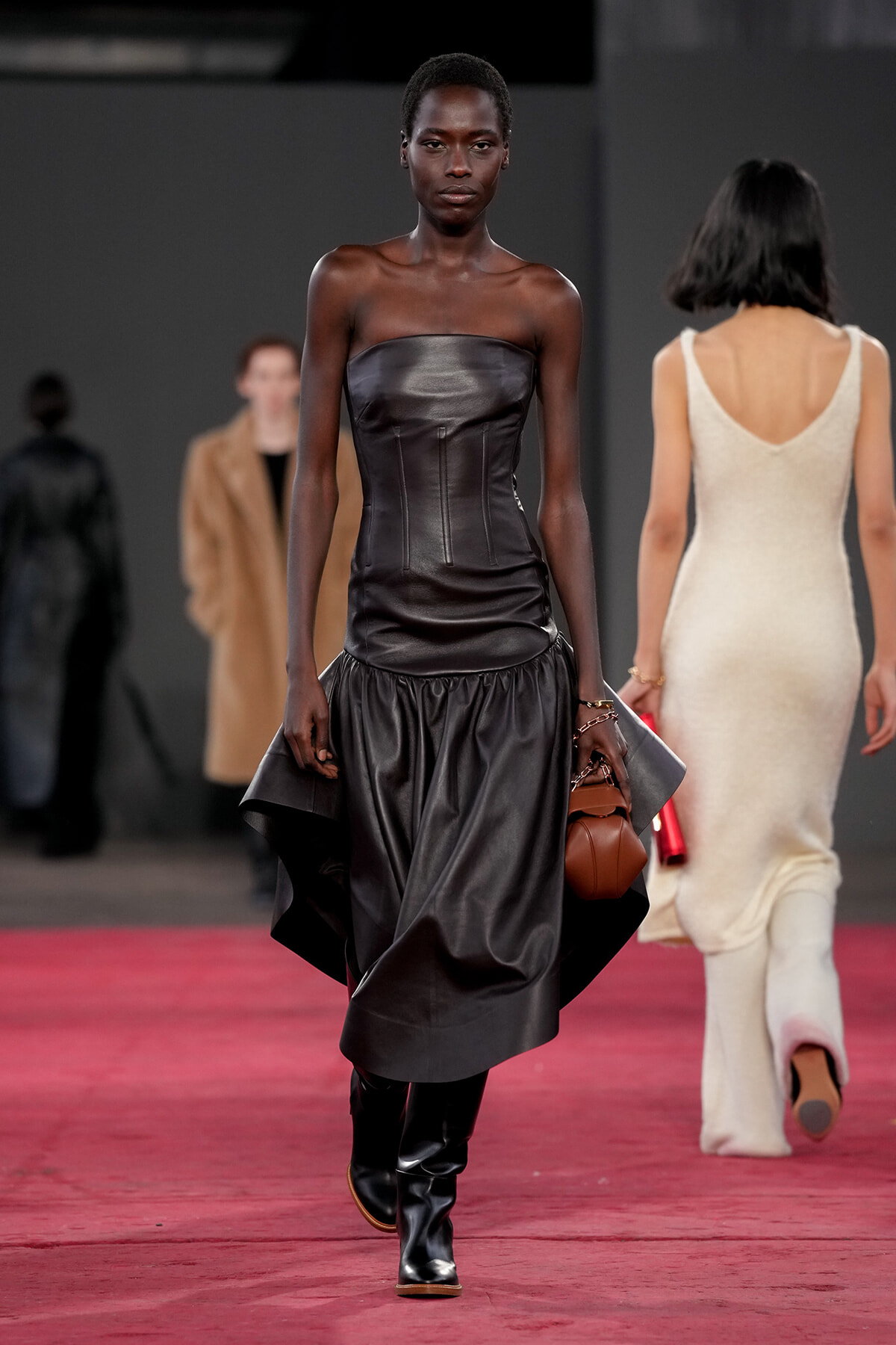Model walking a fashion show wearing a strapless black leather gown with a ruched, voluminous skirt and knee-high boots, carrying a brown handbag.