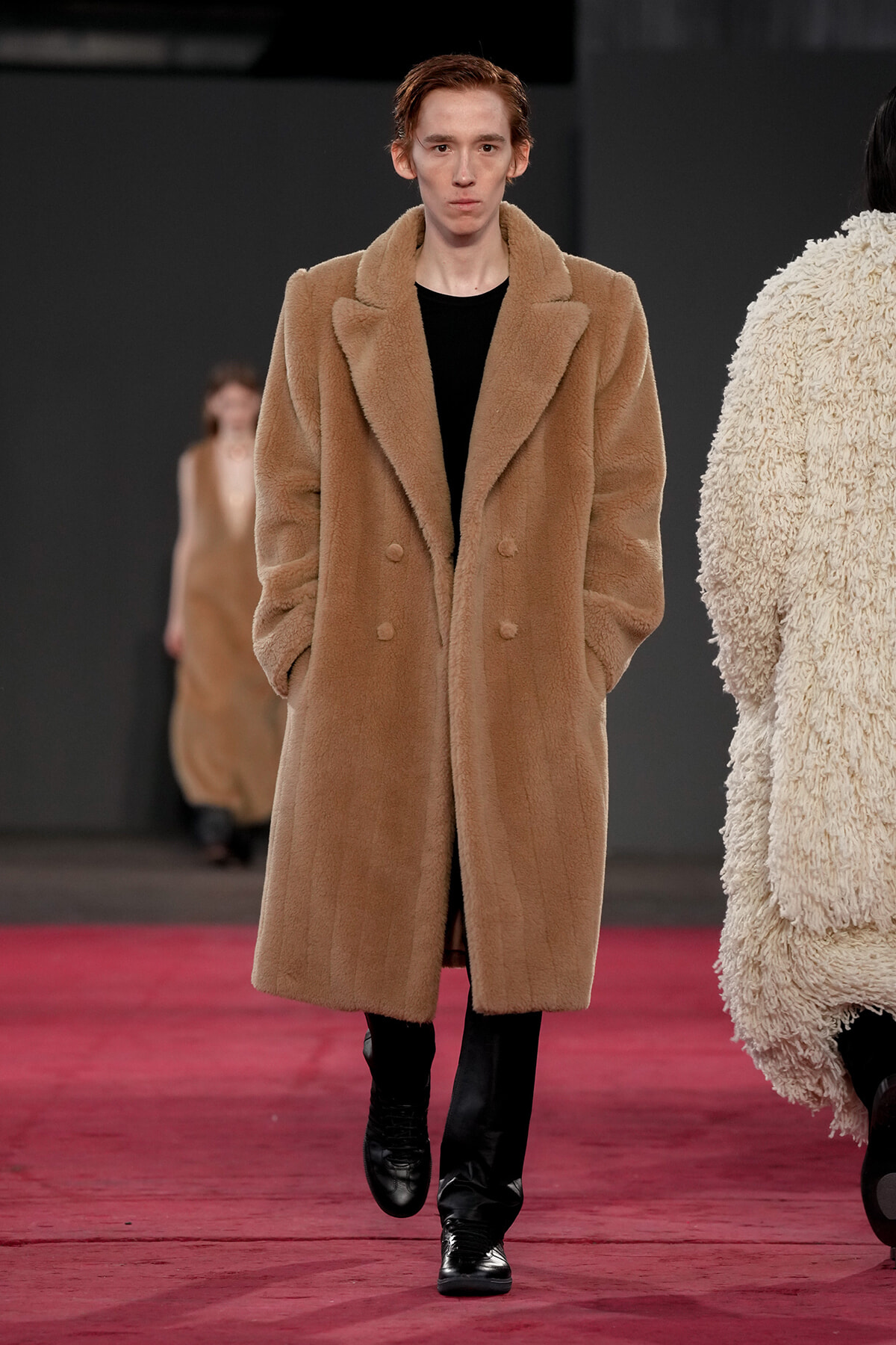 Model walking the runway in a long tan teddy coat over a black top, on a red carpeted catwalk.