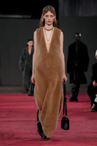 Model walks a runway wearing a tan fuzzy sleeveless dress, black boots, and a small black handbag, against a red runway backdrop.