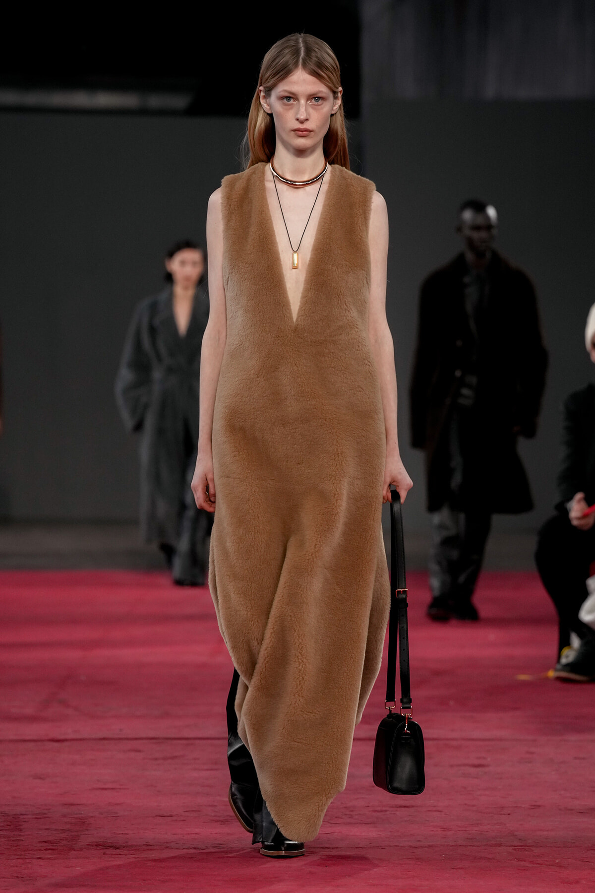 Model walks a runway wearing a tan fuzzy sleeveless dress, black boots, and a small black handbag, against a red runway backdrop.