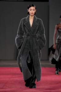Model walking the runway in a long dark gray textured belted coat with oversized sleeves and wide lapels on a red-carpet runway.