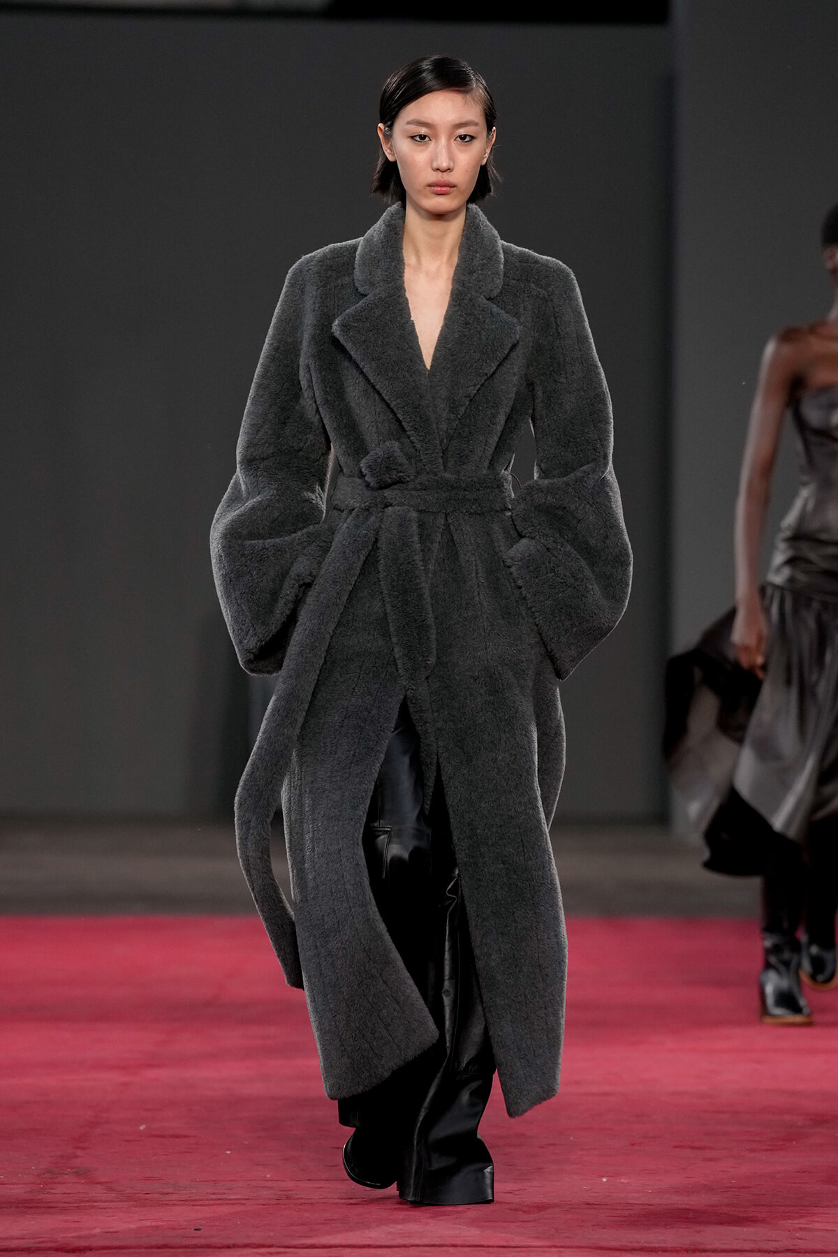 Model walking the runway in a long dark gray textured belted coat with oversized sleeves and wide lapels on a red-carpet runway.