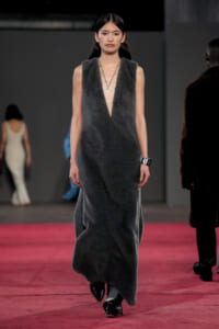 Model walking a fashion show in a sleeveless, deep V, charcoal grey textured dress with a pendant necklace and black footwear on a pink runway surface.