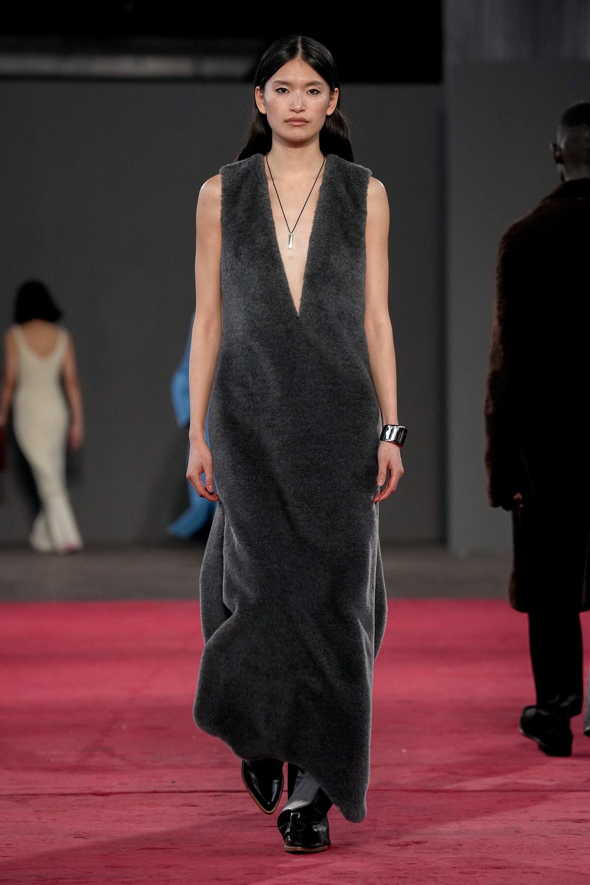 Model walking a fashion show in a sleeveless, deep V, charcoal grey textured dress with a pendant necklace and black footwear on a pink runway surface.