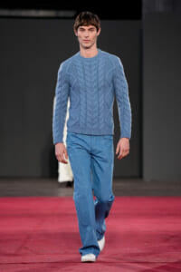 Male model walking a runway in a light blue cable-knit sweater and blue jeans.