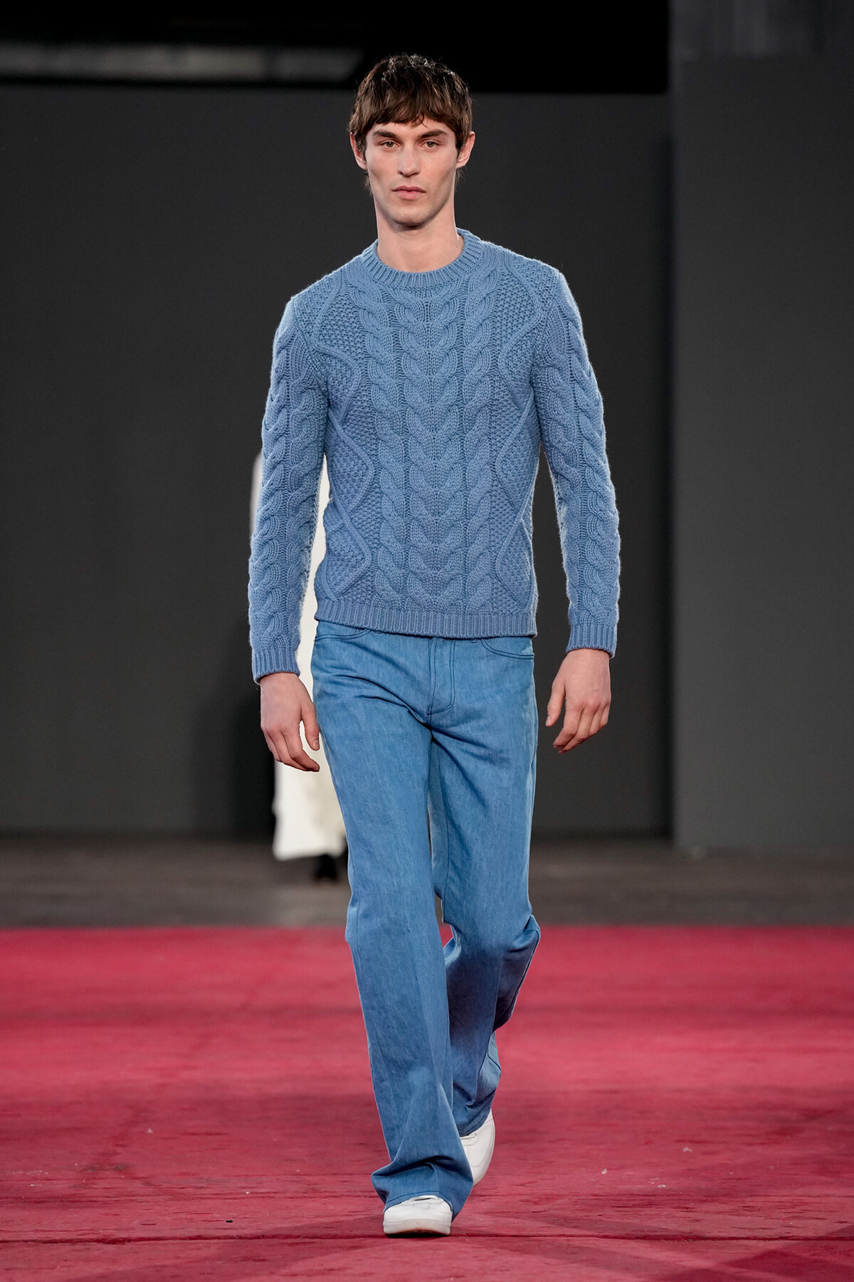 Male model walking a runway in a light blue cable-knit sweater and blue jeans.