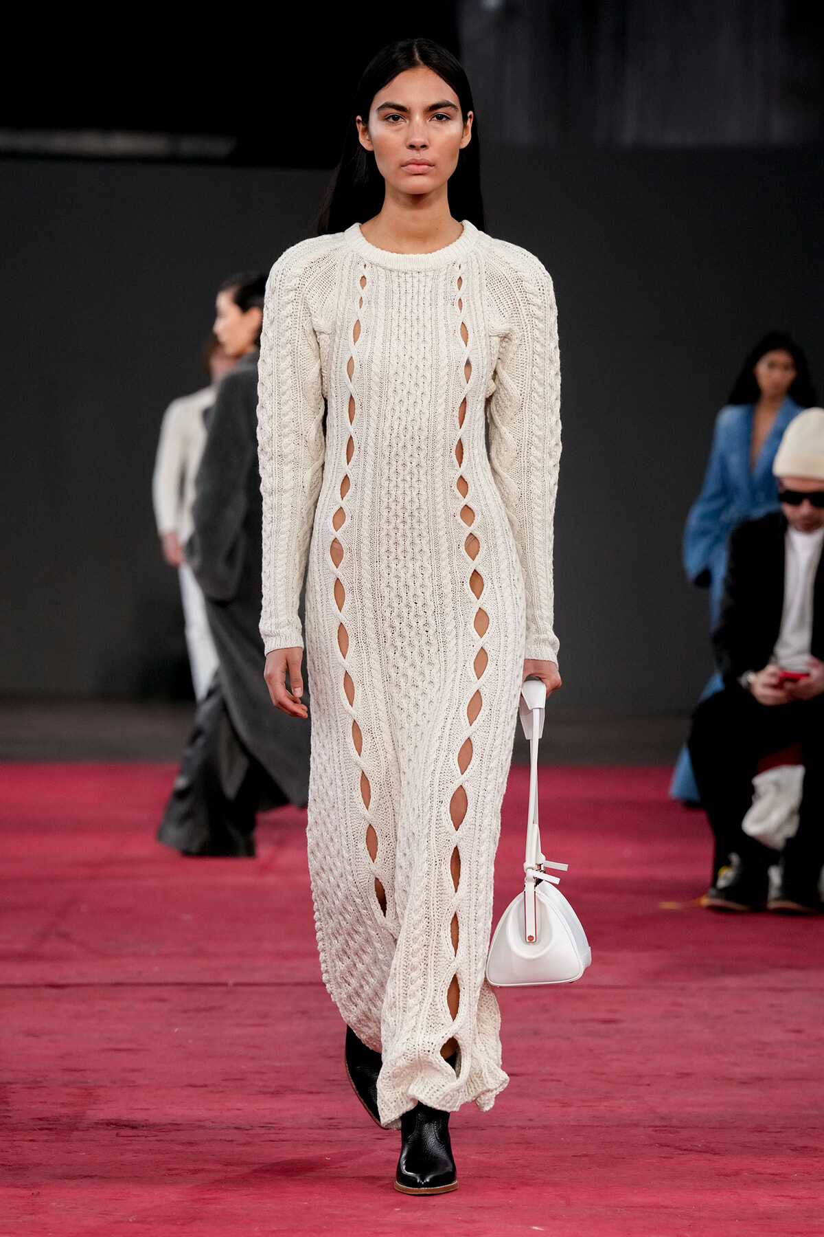 Model walking a runway in a cream knit maxi dress with vertical cutouts, carrying a white bag.