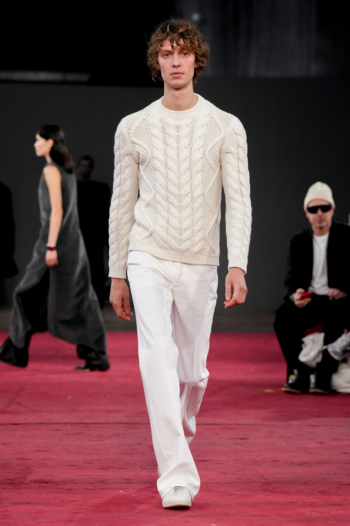 Male model walks the runway in a cream cable-knit sweater and white trousers, on a red-pile runway with spectators in background.