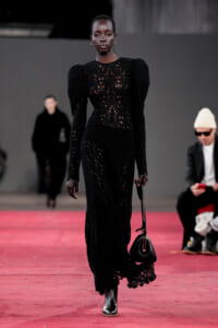 Model walks a red-carpet runway in a long black lace dress, carrying a small black handbag.
