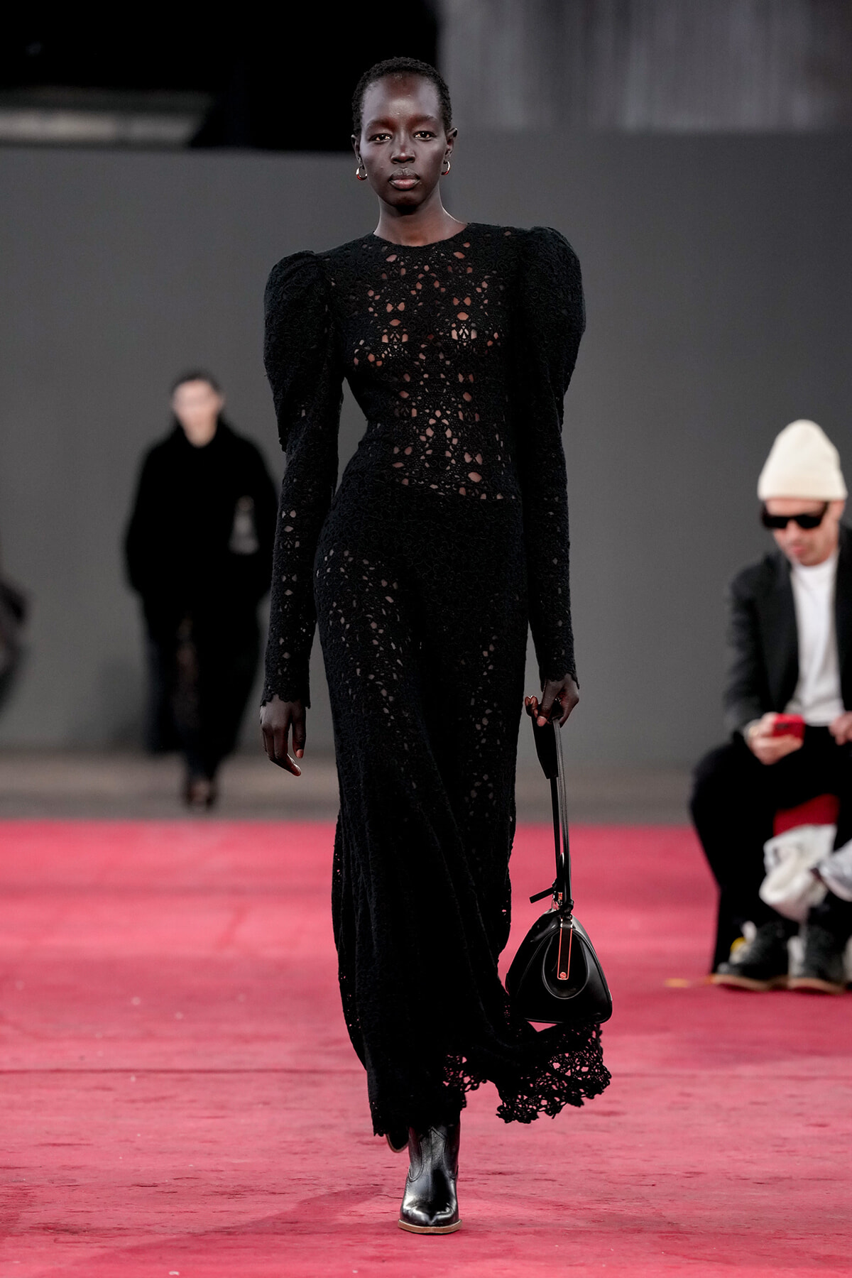 Model walks a red-carpet runway in a long black lace dress, carrying a small black handbag.