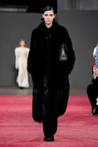 Model walks a fashion show wearing a black fur coat and matching dress, carrying a small black handbag on the runway.