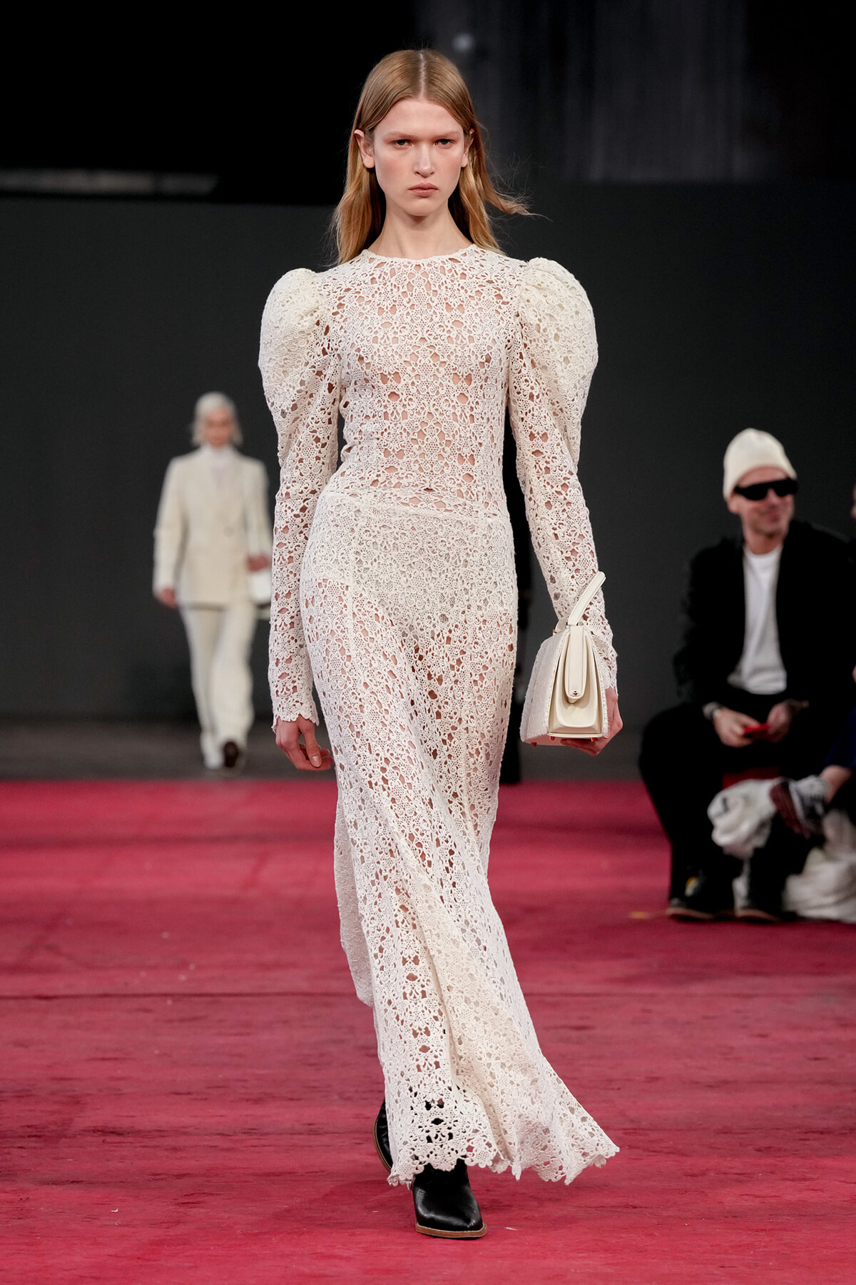 Model walks a runway in a white lace, long-sleeve gown with puff shoulders, holding a small white handbag.