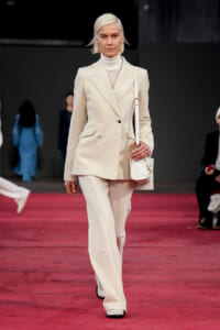 Model wearing a cream double-breasted suit with a lace top, walking a red runway while carrying a white handbag.
