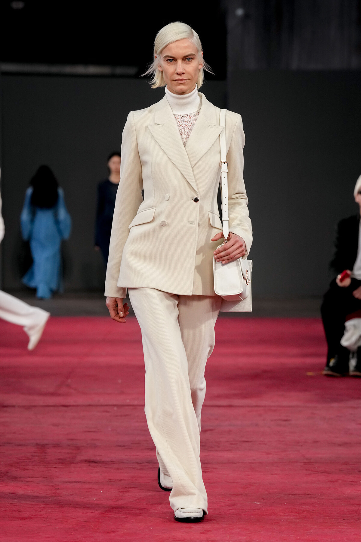 Model wearing a cream double-breasted suit with a lace top, walking a red runway while carrying a white handbag.