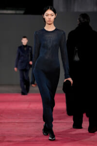 Female model walks a red runway in a dark navy knit full-length dress, with another blurred model in a suit behind her.