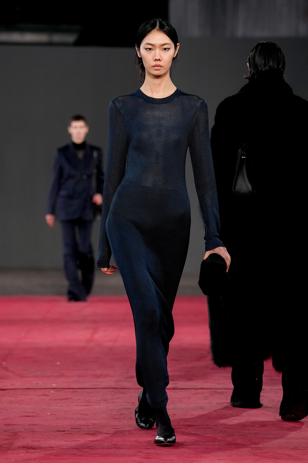 Female model walks a red runway in a dark navy knit full-length dress, with another blurred model in a suit behind her.