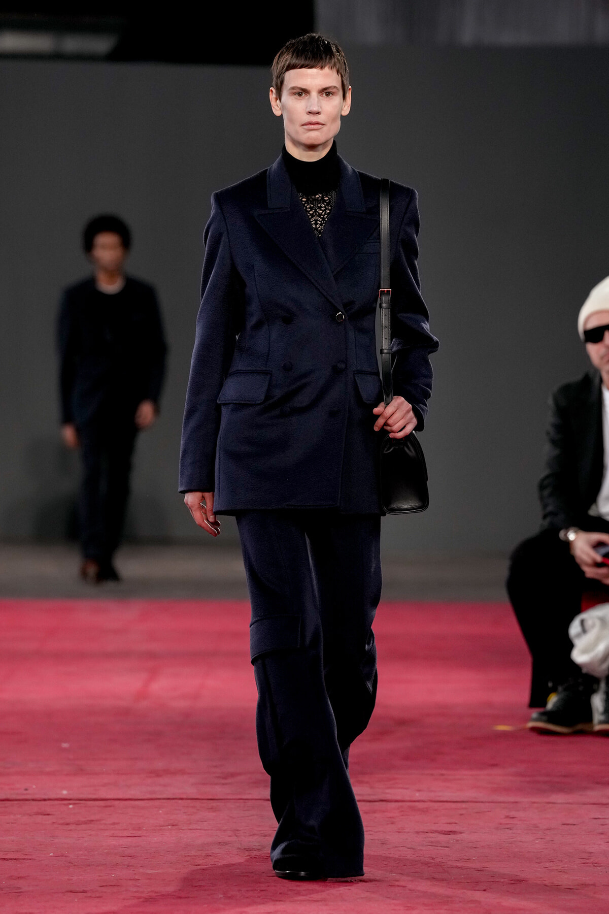 Model walks a navy double-breasted suit with a lace‑top, carrying a black shoulder bag on a red runway.