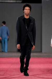Male model walks the runway in a black suit with velvet lapels and a black shirt, mid-stride on a red carpet.