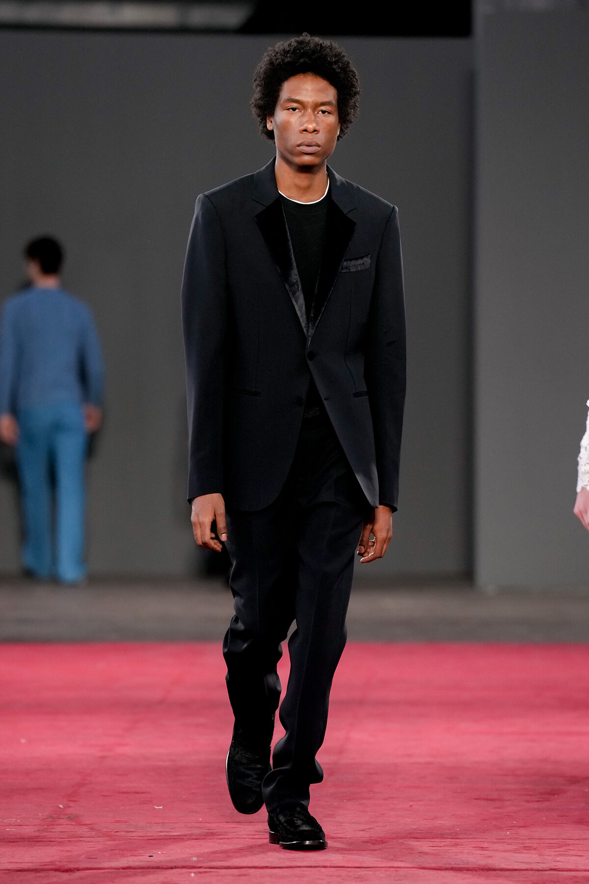 Male model walks the runway in a black suit with velvet lapels and a black shirt, mid-stride on a red carpet.