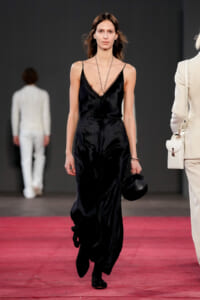 Model on a fashion runway wearing a black satin V-neck slip dress with thin straps, holding a small black handbag.