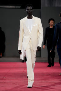 Male model in a cream suit with white shirt walks the pink runway at a fashion show.