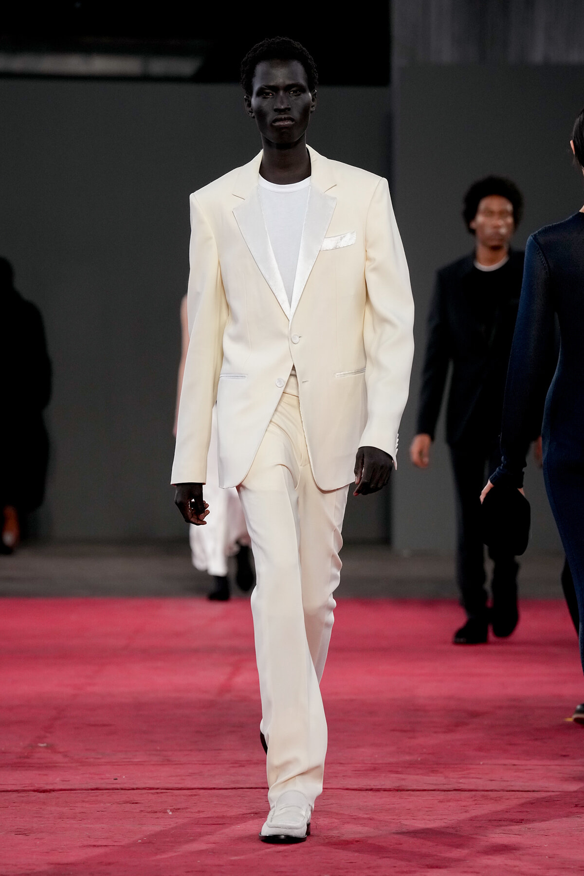 Male model in a cream suit with white shirt walks the pink runway at a fashion show.