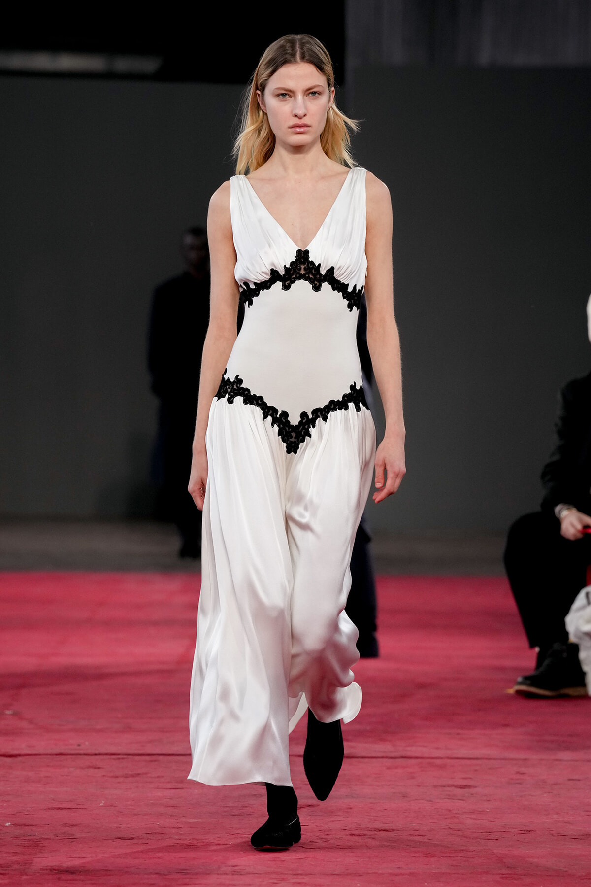 Model walking the runway in a white satin gown with black lace trim at the bodice and waist.