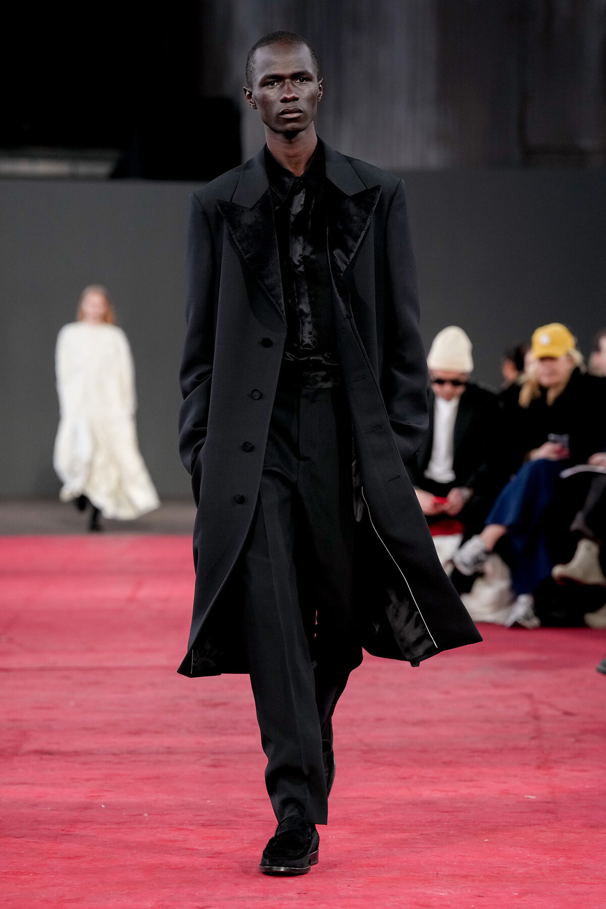 Model walking down a red runway in a long black suit with a velvet lapel, focused expression, hands in pockets, audience in background.