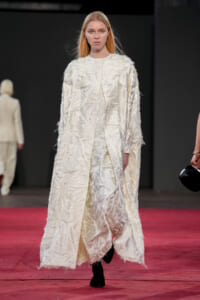 Model walks the runway in a long textured ivory coat with fringe, on a red carpeted catwalk.