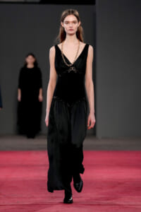 Model walks the runway wearing a black sleeveless gown with lace details and a chunky gold chain necklace against a dark backdrop.