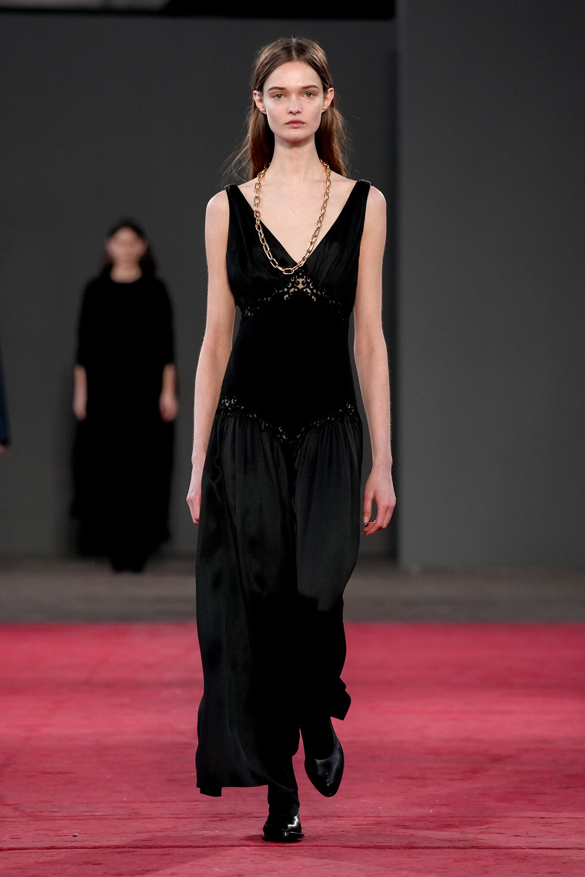 Model walks the runway wearing a black sleeveless gown with lace details and a chunky gold chain necklace against a dark backdrop.